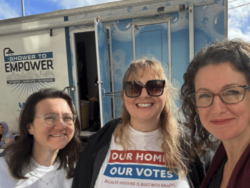Three female presenting people outside, smiling for the camera, wearing shirts with Our Homes Our Votes logos.