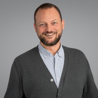 Portrait of David Gonzalez Rice, smiling, wearing a grey sweater over a light dress shirt.