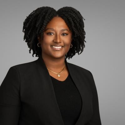 Portrait of Zenayah Roache, smiling, with medium length, curly brown hair, wearing a dark jacket.